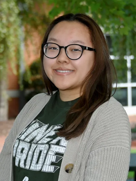 East Asian woman with black reading glasses, long straight brown hair wearing a niner pride green shirt and a beige sweater