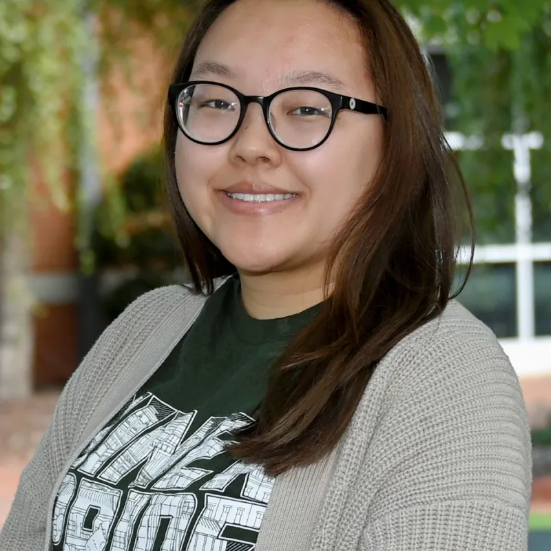 East Asian woman with black reading glasses, long straight brown hair wearing a niner pride green shirt and a beige sweater