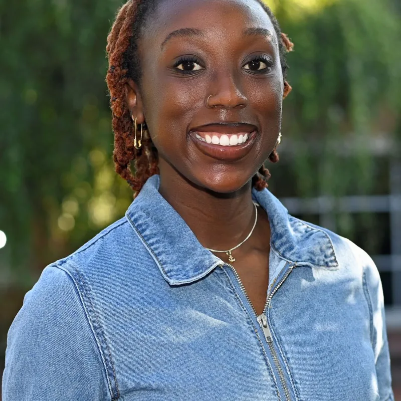 portrait of young woman smiling with dark skin, shoulder length bronze locks, and a light blue jean shirt 